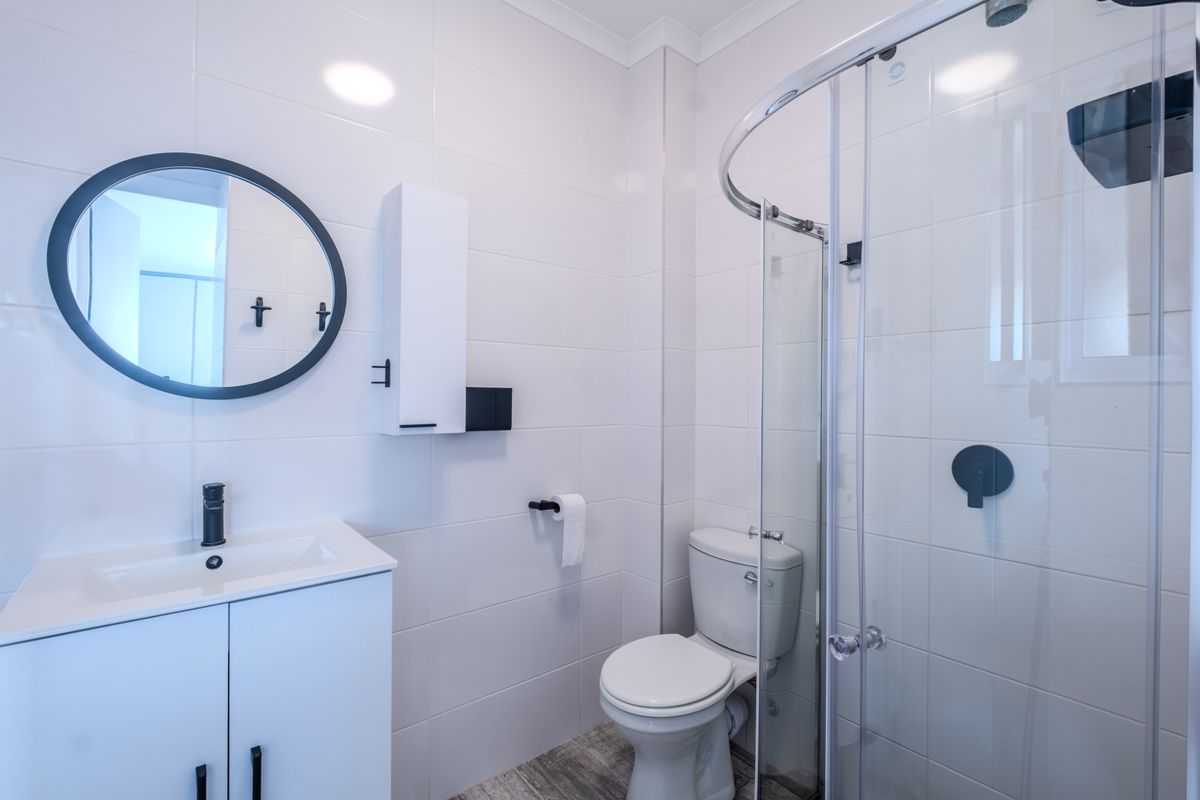A modern, clean bathroom featuring a white vanity, round mirror, toilet, and a glass-enclosed shower with contemporary black fixtures.
