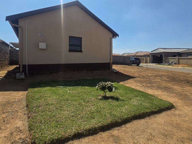Rear view of the property featuring a plastered exterior wall, tiled roof, and a small patch of lawn within an unlandscaped yard.
