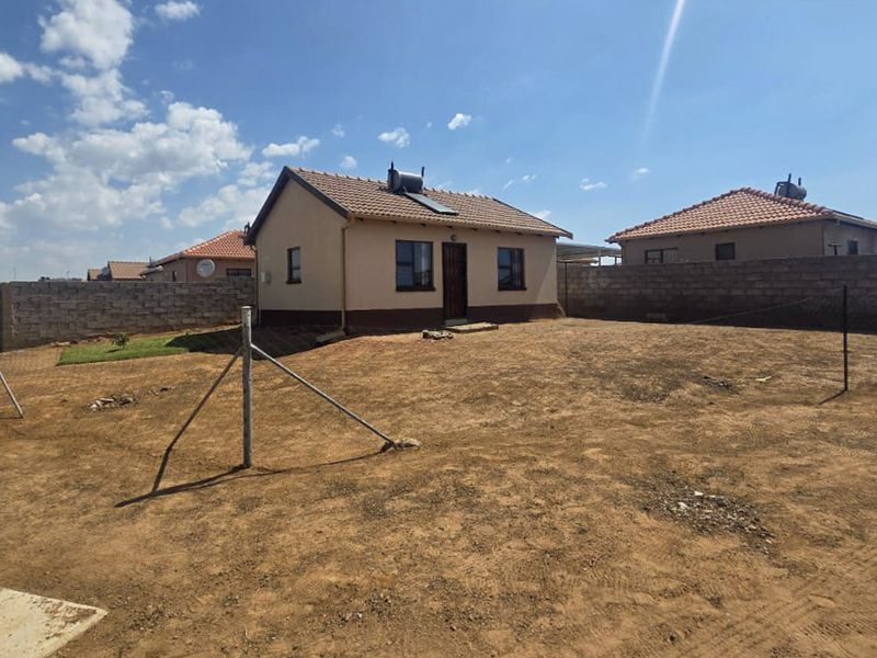 Rear view of a detached house featuring a tiled roof, solar water heater, and a large, undeveloped yard enclosed by perimeter walls.
