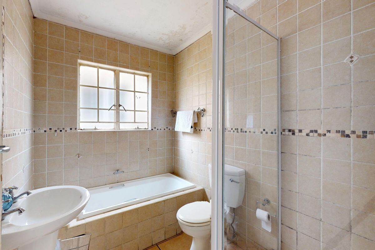A dated bathroom featuring a built-in bathtub, pedestal basin, toilet, and glass shower enclosure, with visible ceiling water damage requiring maintenance.