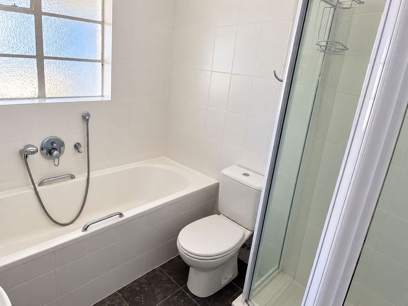 A clean, functional bathroom featuring a bathtub, separate shower enclosure, and toilet with neutral wall tiling and dark floor tiles.