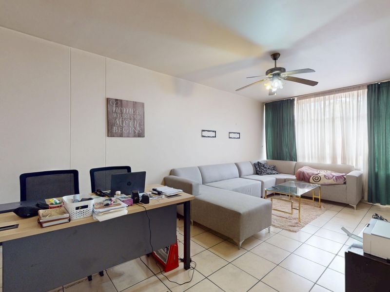 An open-plan living area featuring tiled flooring, a ceiling fan, and large windows, providing a versatile space for both relaxation and home office use.