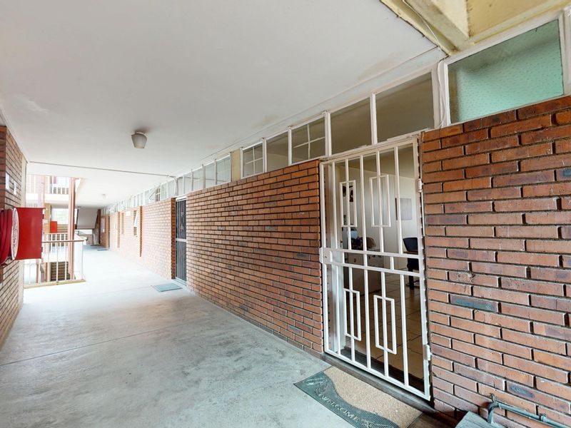 An exterior apartment building corridor featuring exposed brick walls, a security gate entrance, and a wall-mounted fire hose reel.