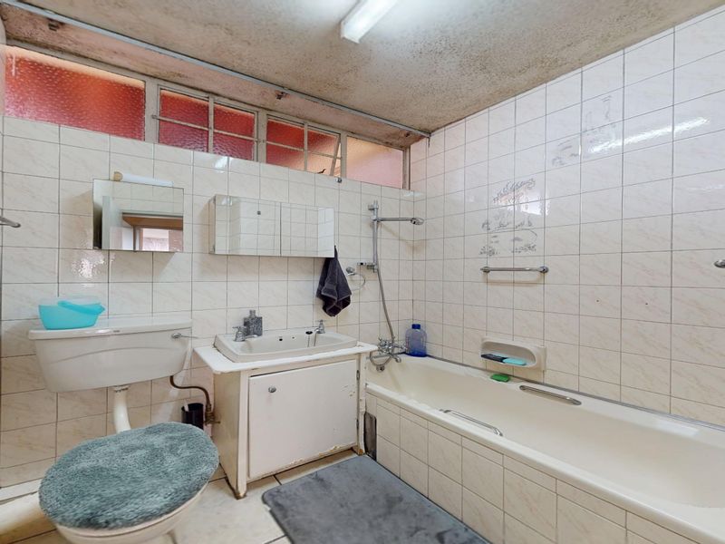 A dated bathroom featuring a bathtub, shower attachment, wall-mounted basin with vanity, and toilet, finished with full-height wall tiling and high-level windows.