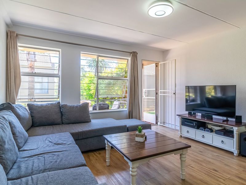 A bright living room featuring laminate flooring, large windows with natural light, and direct access to an outdoor area through a security gate.