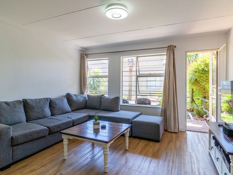A bright and well-maintained living room featuring laminate flooring, large windows with security bars, and direct access to the garden area.