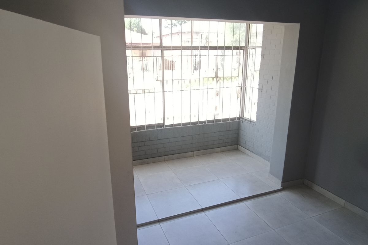 An empty living area featuring neutral tiled flooring, grey painted walls, and a large window fitted with security bars.