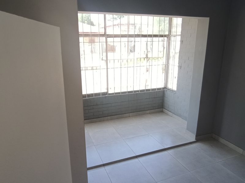 An empty living area featuring neutral tiled flooring, grey painted walls, and a large window fitted with security bars.