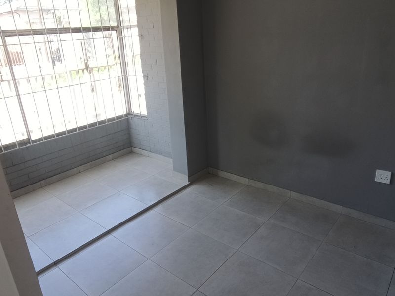 An empty bedroom featuring tiled flooring, grey painted walls with visible staining, and large windows fitted with security bars.