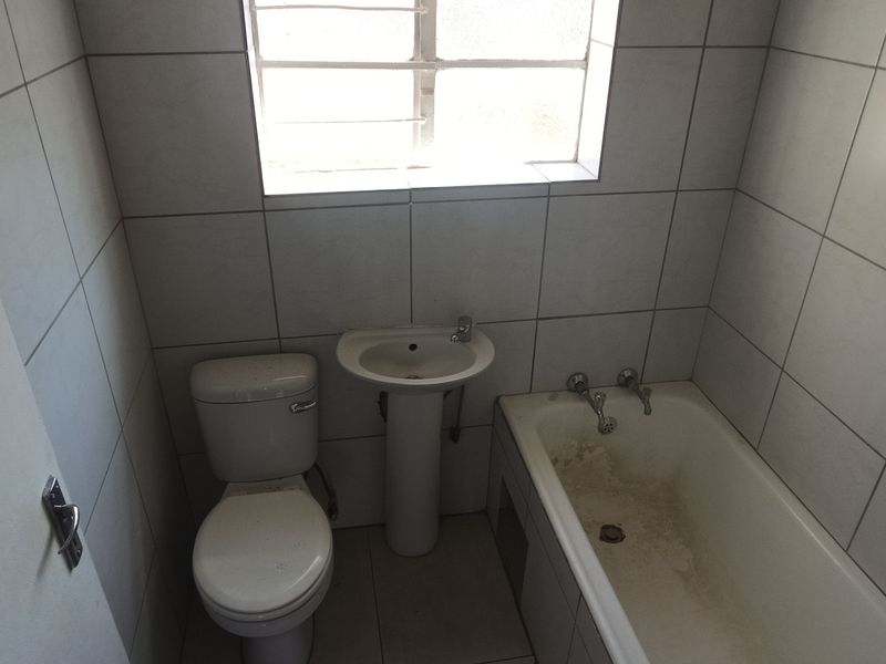 A compact bathroom featuring a pedestal basin, toilet, and bathtub with full-height wall tiling and a window with security bars.