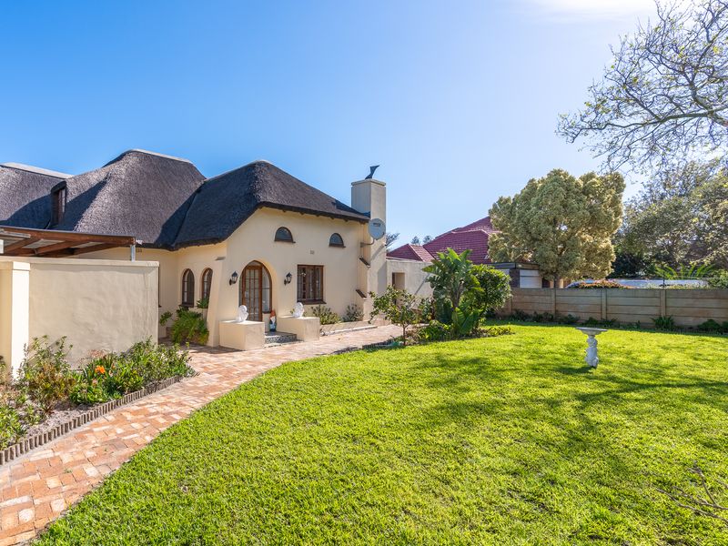 A well-maintained rear garden featuring a charming thatched-roof house, a brick-paved pathway, and a spacious lawn area.