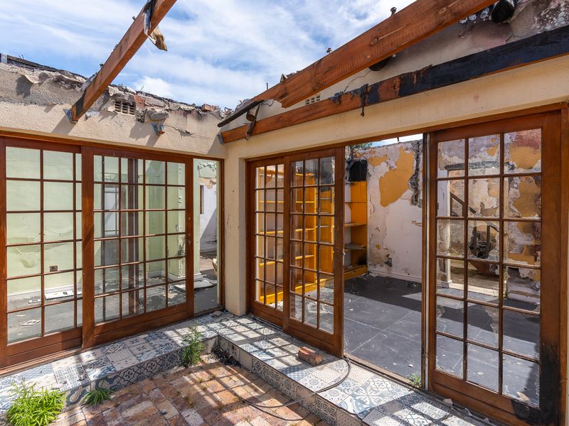 A severely fire-damaged patio area showing exposed roof rafters, damaged wall finishes, and timber sliding doors leading into a ruined interior space.