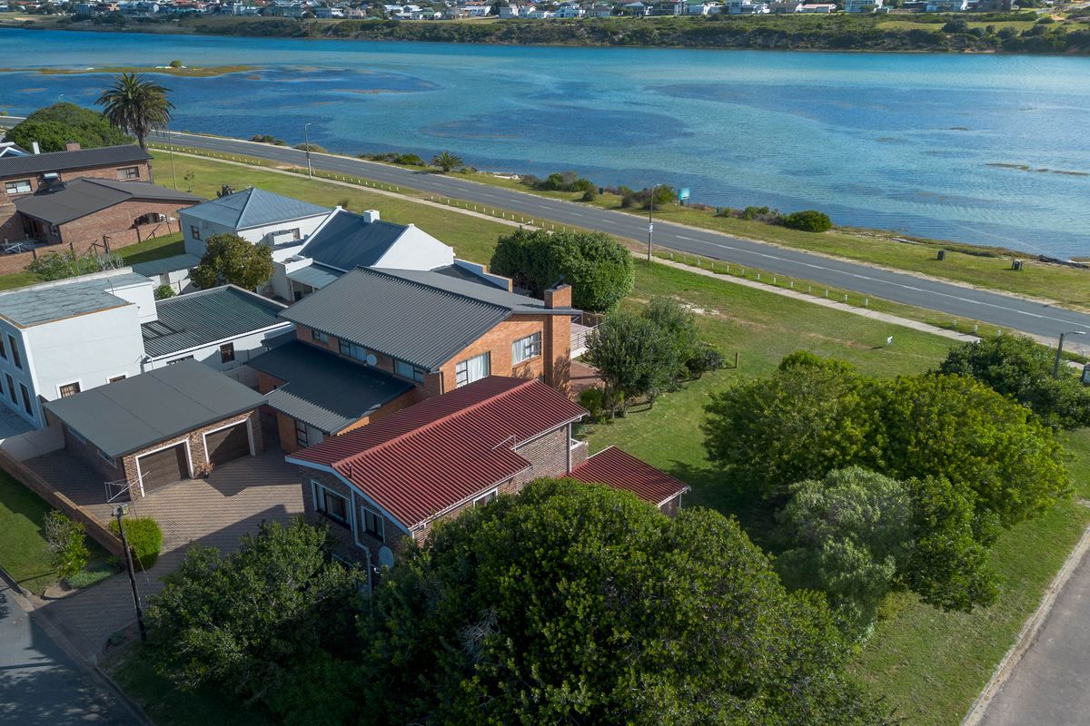 Aerial view of a brick house with a metal roof, double garage, and paved driveway, situated near a coastal road and water.