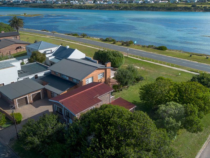 Aerial view of a brick house with a metal roof, double garage, and paved driveway, situated near a coastal road and water.