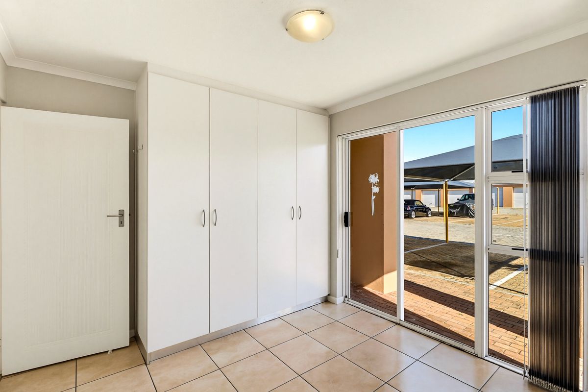 A bright, empty bedroom featuring built-in cupboards, tiled flooring, and a sliding glass door providing direct access to the exterior parking area.