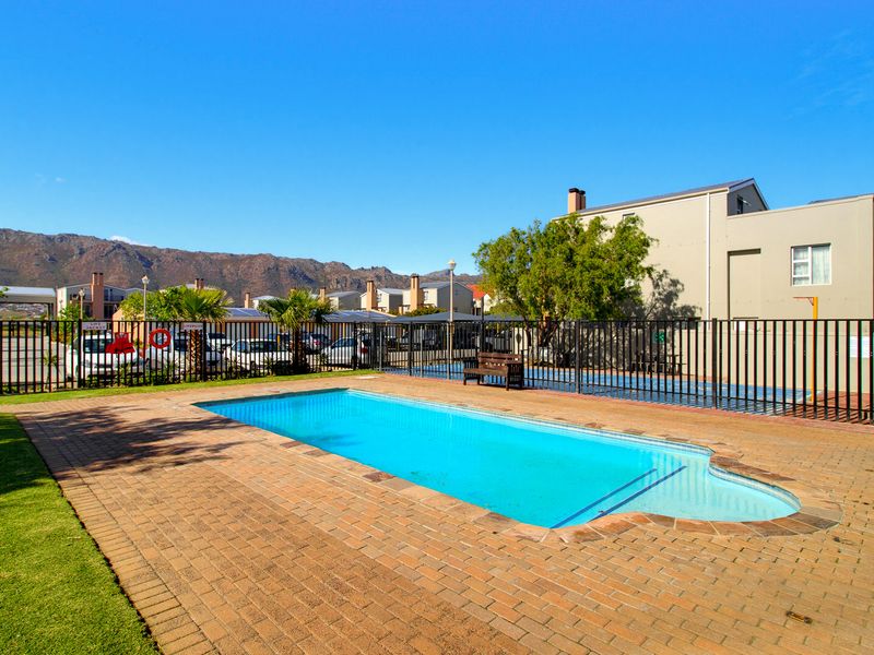 A well-maintained communal swimming pool area featuring brick paving, secure metal perimeter fencing, and landscaped surroundings with mountain views.
