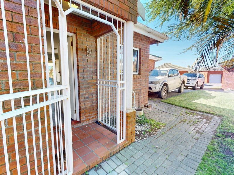 Front entrance of the townhouse featuring a brick facade, security gate, tiled porch, and paved driveway area.