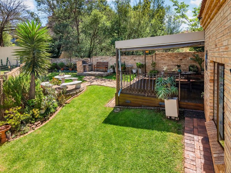 A well-maintained rear garden featuring a raised wooden deck with a retractable awning, a built-in braai area, and established landscaping.