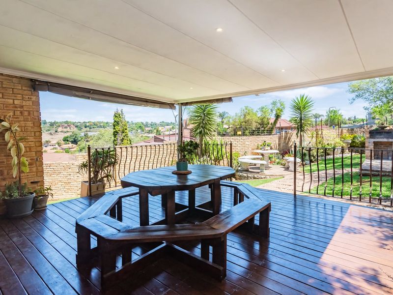 Spacious covered timber deck patio featuring recessed lighting, a brick feature wall, and wrought iron balustrades, overlooking a landscaped garden and scenic views.