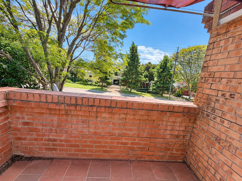 An open-air balcony featuring exposed brick walls and tiled flooring, offering a view of the surrounding neighborhood and mature trees.