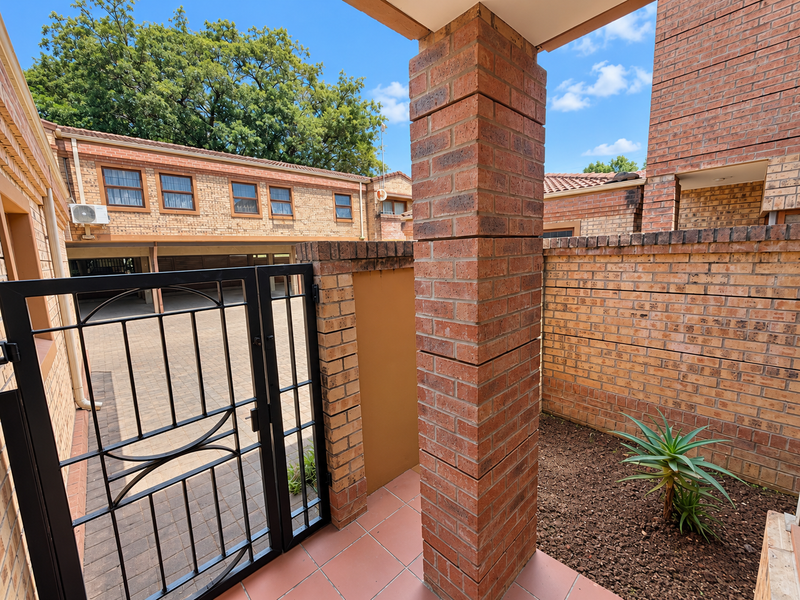 A view of the townhouse entrance featuring a brick pillar, tiled patio area, and a secure metal gate leading to the paved driveway.