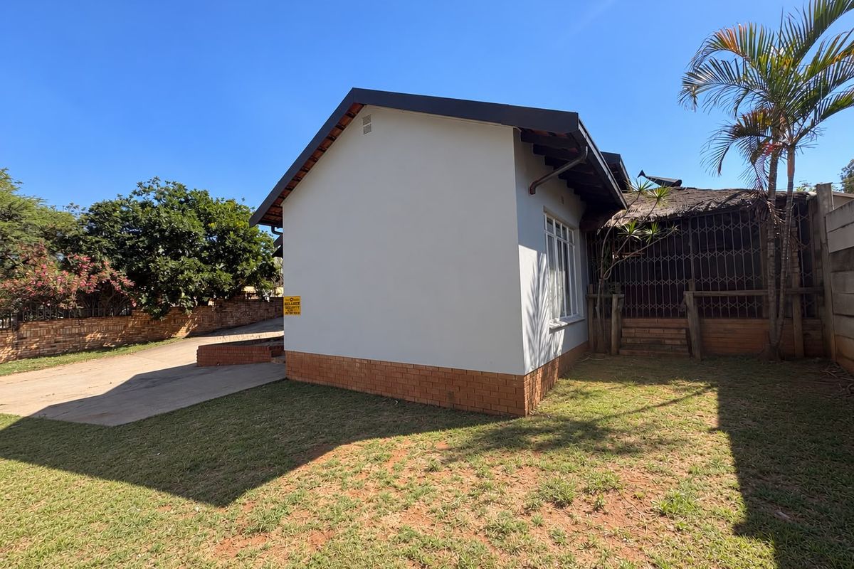 Rear view of the property featuring a brick plinth, metal security gate, and a section of thatched roofing with an adjacent concrete driveway.