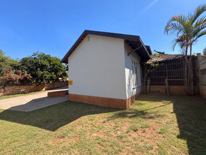 Rear view of the property featuring a brick plinth, metal security gate, and a section of thatched roofing with an adjacent concrete driveway.
