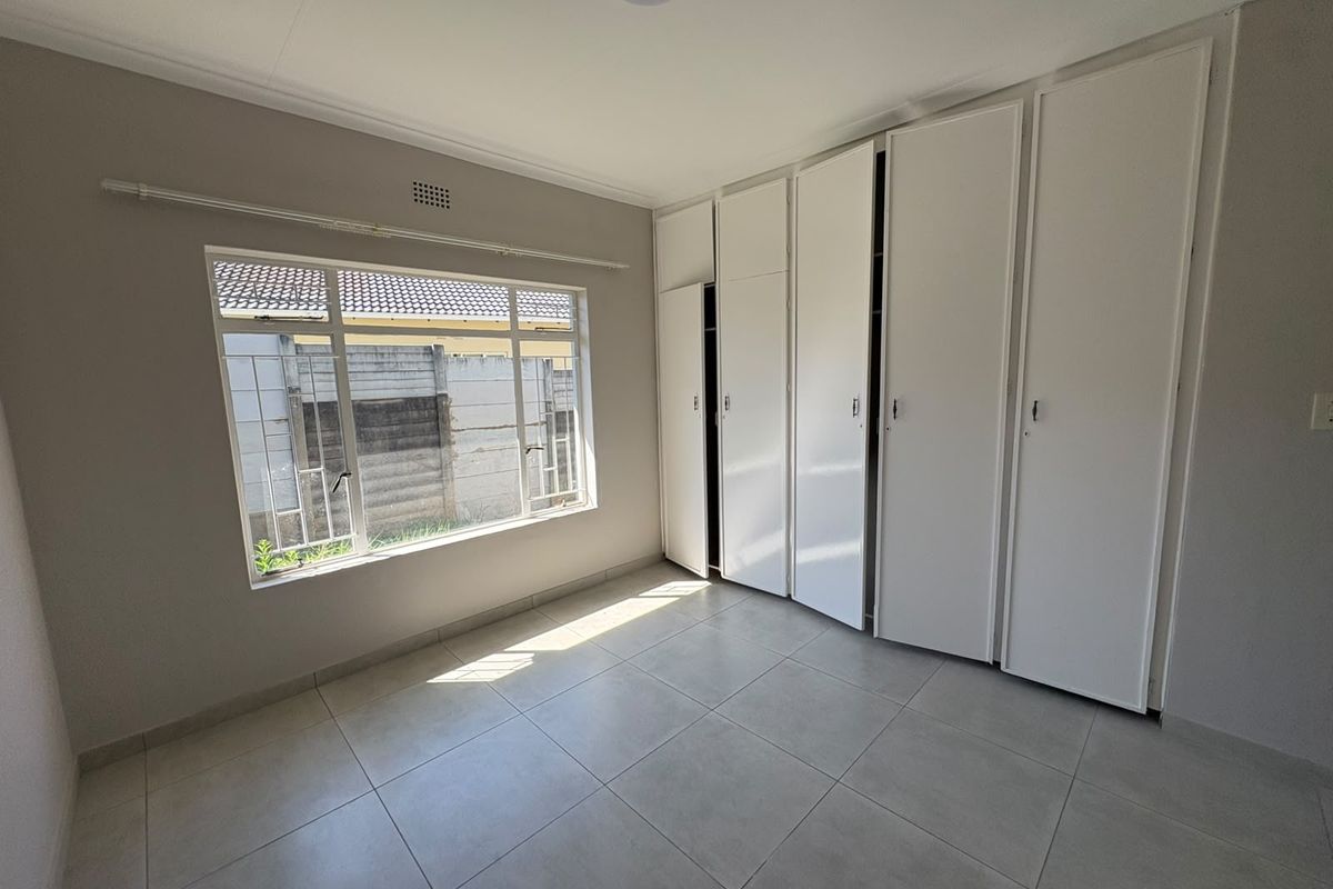 A bright, empty bedroom featuring neutral tiled flooring, built-in white cupboards, and large windows with security bars.