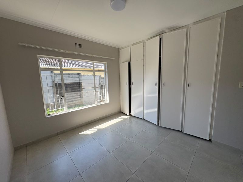 A bright, empty bedroom featuring neutral tiled flooring, built-in white cupboards, and large windows with security bars.