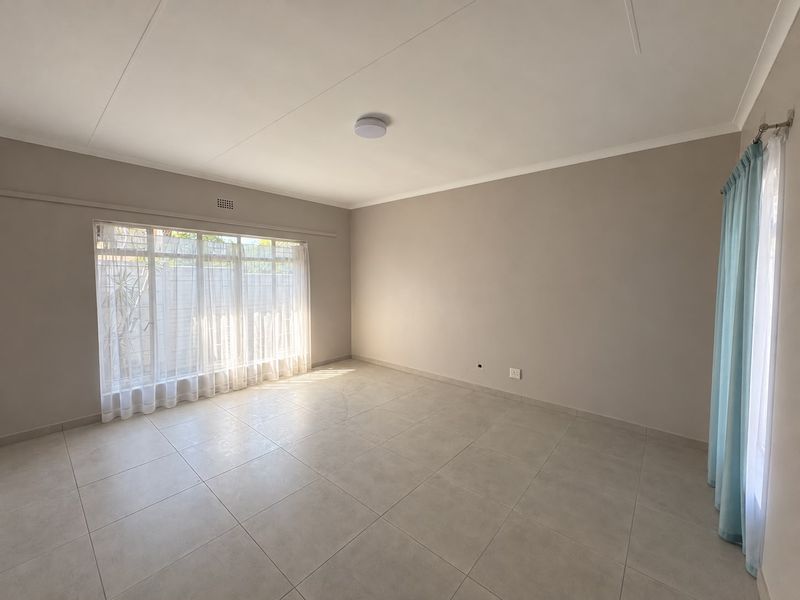 A bright and spacious empty living room featuring neutral tiled flooring, large windows with natural light, and a clean, well-maintained finish.