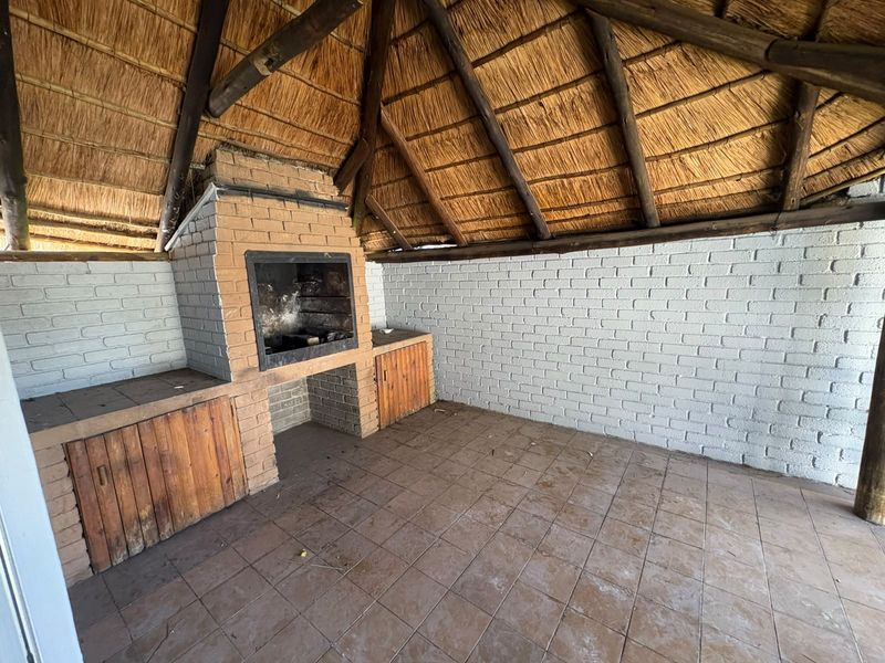 Covered outdoor patio area featuring a traditional thatched roof, exposed timber rafters, and a built-in brick braai with integrated storage cabinetry.