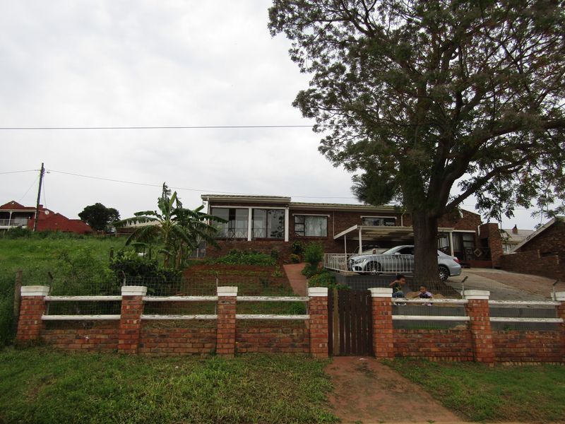 Front view of a single-story brick house featuring a gated entrance, paved driveway, carport, and established garden with a large mature tree.