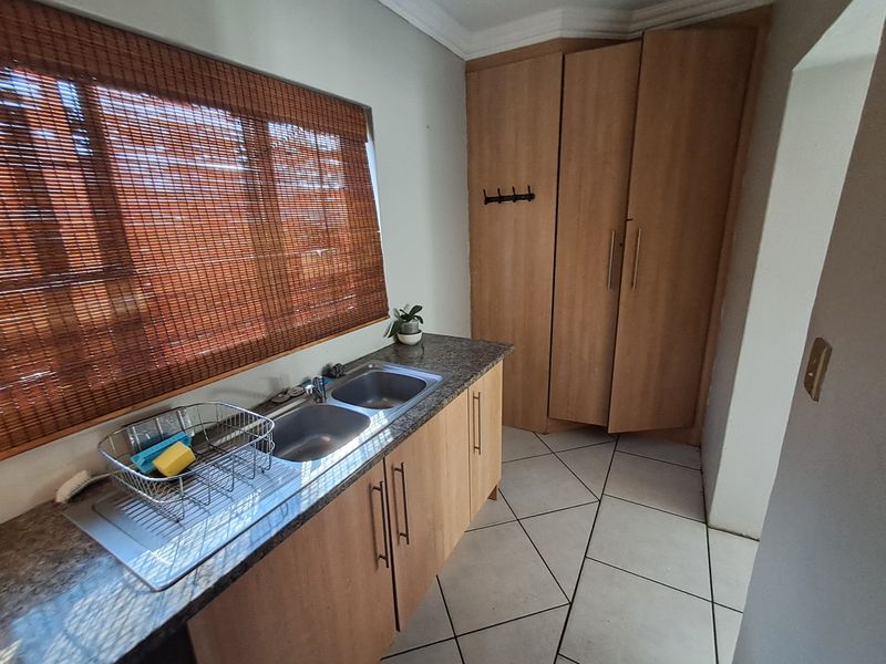 A functional scullery area featuring a double bowl stainless steel sink, granite countertops, laminate cabinetry, and built-in storage cupboards with tiled flooring.
