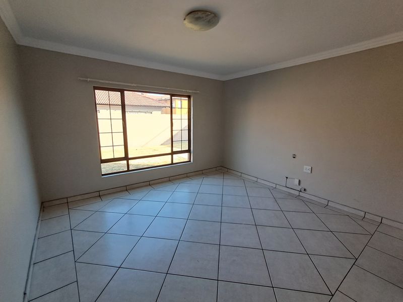 A bright, empty living room featuring neutral wall tones, large window for natural light, and clean tiled flooring throughout.