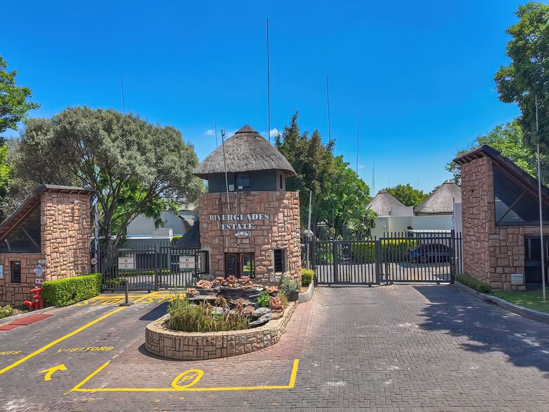 The secure entrance gate of Riverglades Estate featuring stone-clad pillars, a thatched guard house, and a paved driveway with a central water feature.