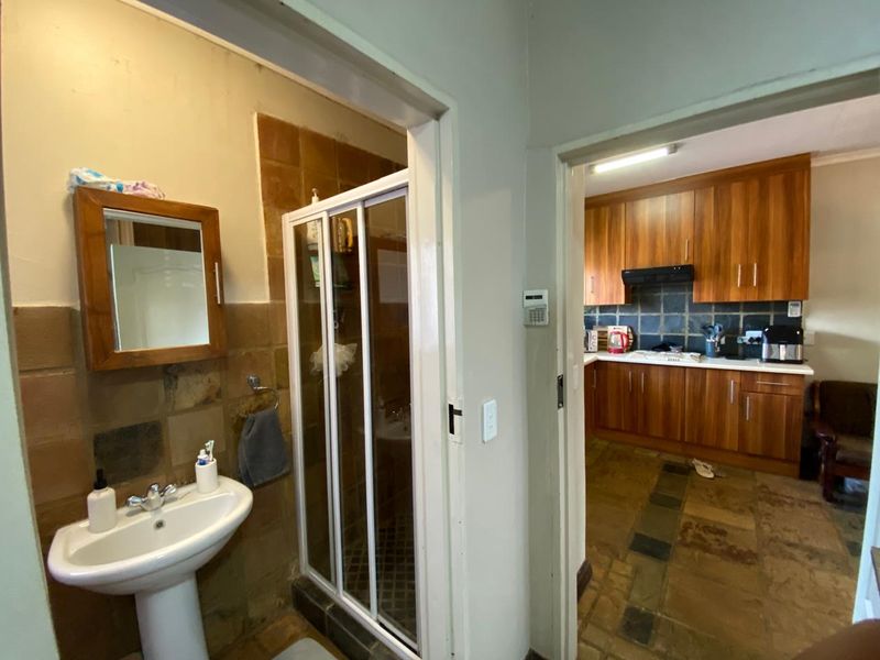 A hallway view connecting a bathroom with a pedestal basin and shower to a kitchen featuring wooden cabinetry and stone flooring.