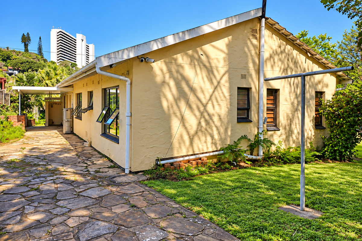 Rear view of the property featuring a stone paved driveway, established lawn, and exterior wall with security cameras and casement windows.