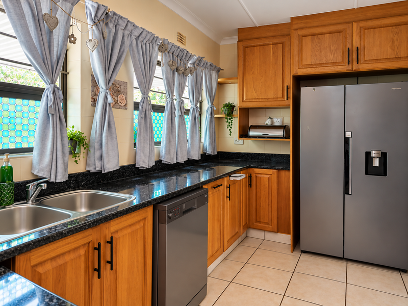 A functional kitchen featuring warm-toned wooden cabinetry, dark granite countertops, a double stainless steel sink, and tiled flooring with integrated dishwasher.