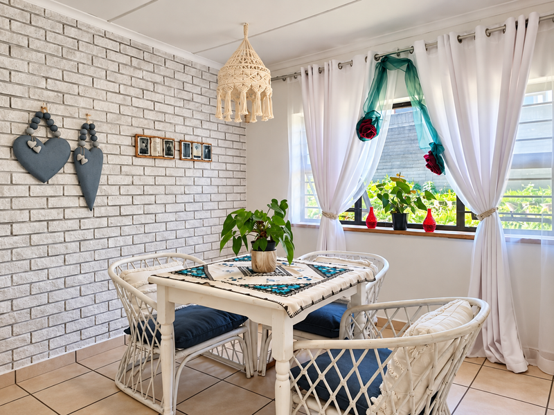 A bright dining area featuring a textured white exposed brick wall, tiled flooring, and a window with security bars overlooking the garden.