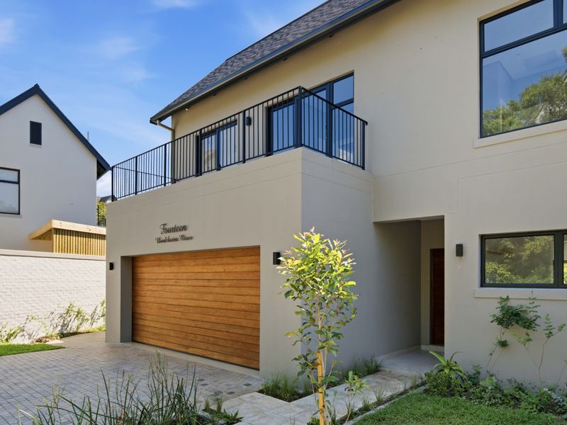 Modern double-storey home featuring a timber garage door, paved driveway, and an upper-level balcony with sleek metal balustrades.