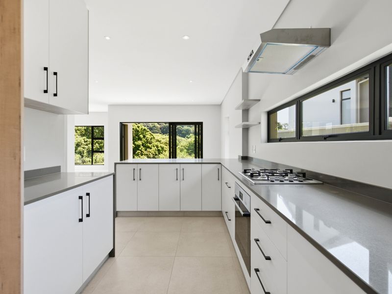 Modern, minimalist kitchen featuring sleek white cabinetry, stone countertops, integrated gas hob, oven, and extractor fan, with large windows overlooking greenery.