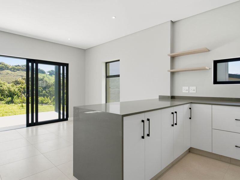 Modern, minimalist kitchen featuring sleek white cabinetry, grey quartz countertops, floating shelves, and large sliding doors opening to a scenic outdoor view.