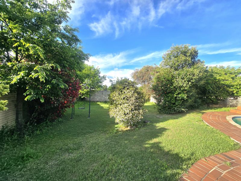 A spacious backyard garden featuring a well-maintained lawn, mature trees, perimeter boundary walls, and a brick-paved pool surround.