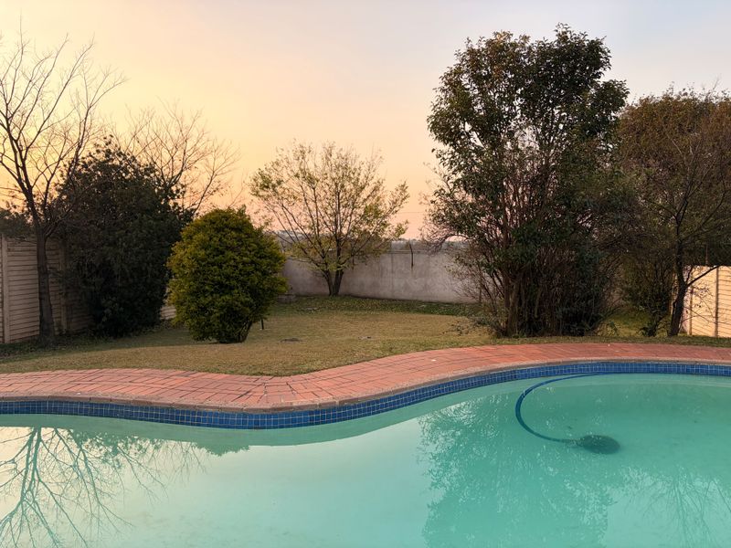 A backyard pool area featuring a curved swimming pool with brick coping, surrounded by a lawn and perimeter boundary wall.