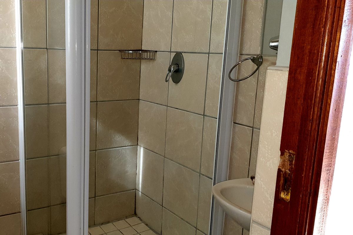 A compact bathroom featuring a tiled shower enclosure, wall-mounted basin, and neutral tiling. The door frame shows visible wear and requires maintenance.