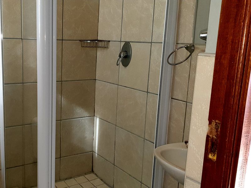 A compact bathroom featuring a tiled shower enclosure, wall-mounted basin, and neutral tiling. The door frame shows visible wear and requires maintenance.