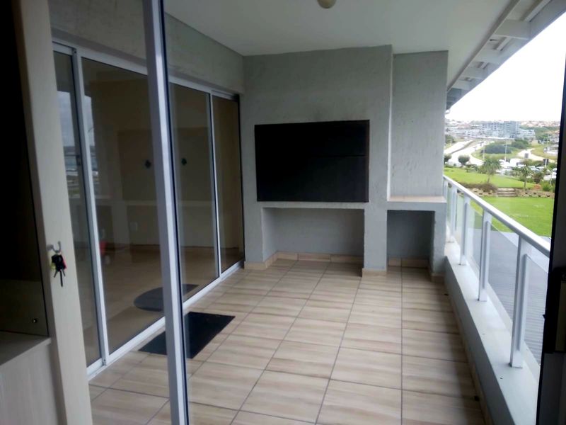 A spacious covered balcony featuring tiled flooring, a built-in braai, and a modern glass balustrade with views over the surrounding area.