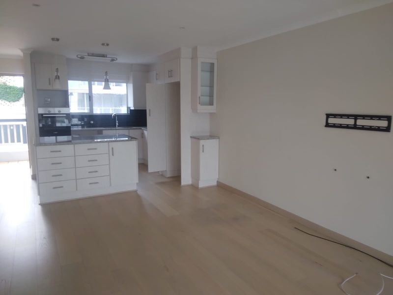 An open-plan living and kitchen area featuring modern white cabinetry, granite countertops, built-in oven, and light hardwood flooring in a clean, vacant space.