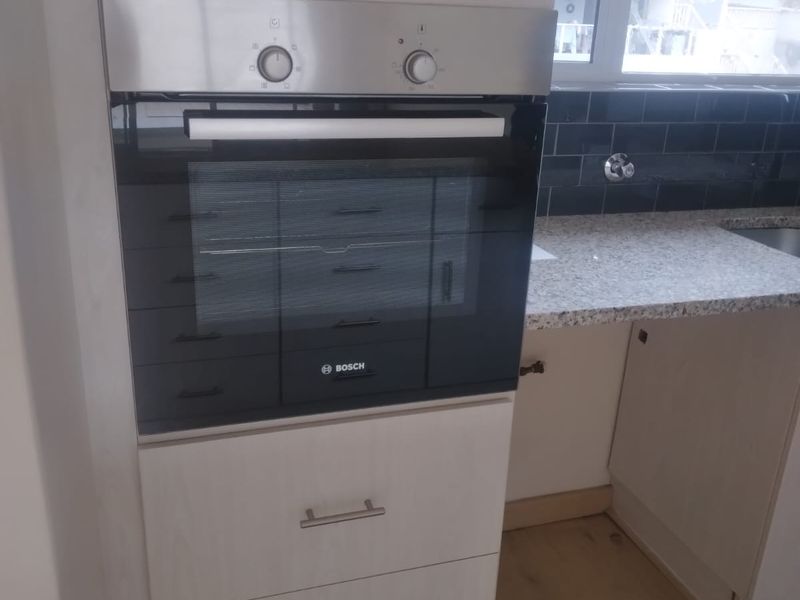 A modern kitchen area featuring a built-in Bosch oven, granite countertops, and a stylish dark subway tile backsplash with light wood-finish cabinetry.