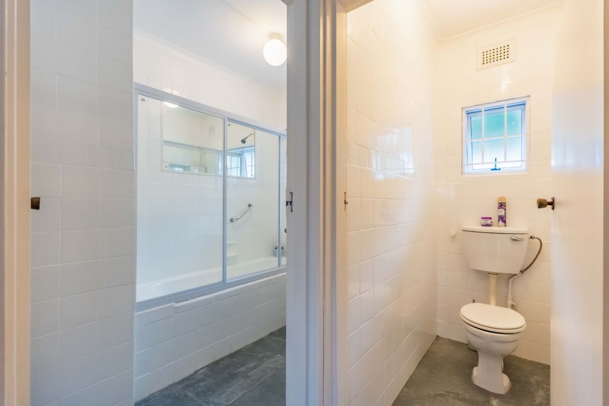 A functional bathroom featuring a bathtub with shower screen and a separate toilet area, finished with white wall tiles and grey flooring.
